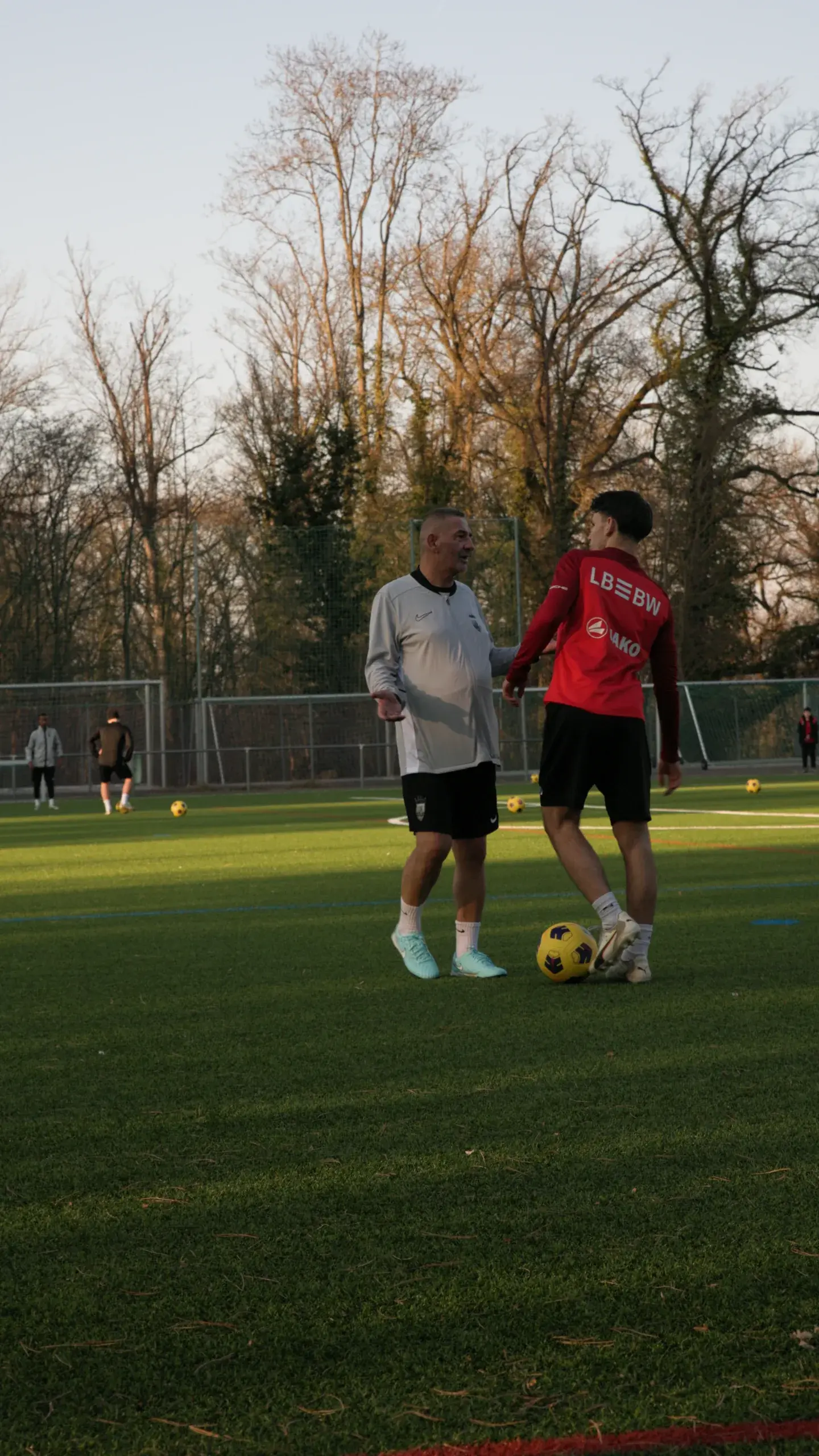 Trainer im Einzeltraining mit einem Spieler, Ball zwischen beiden, Abendlicht auf dem Platz